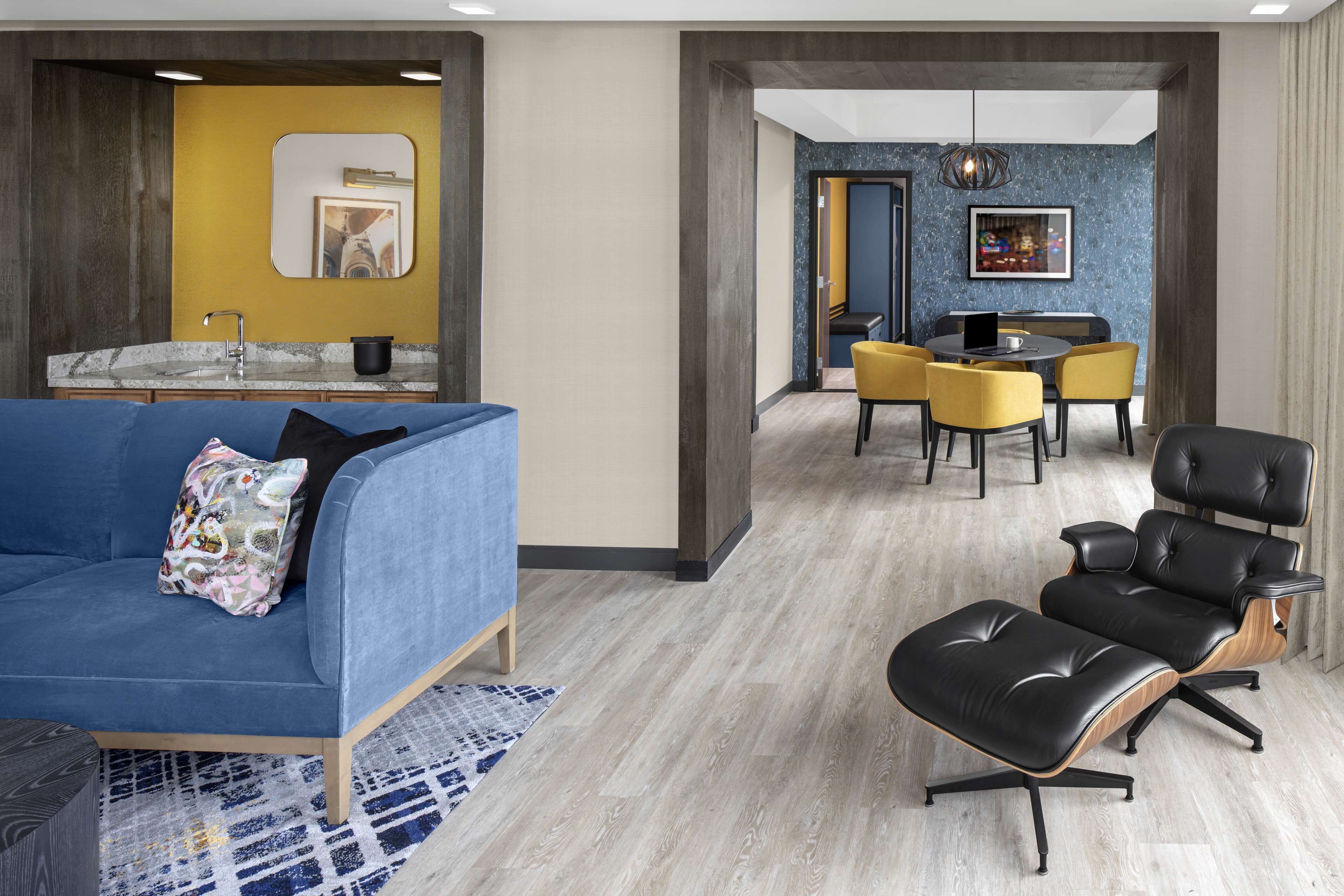 A modern living room with a blue sofa, a black leather chair, and a dining area with yellow chairs.