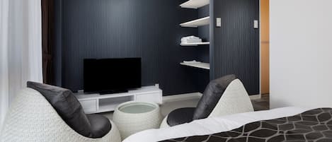 A modern hotel room with a flat-screen TV on a white console, a bed with a black and white patterned bedspread, and two grey upholstered chairs.