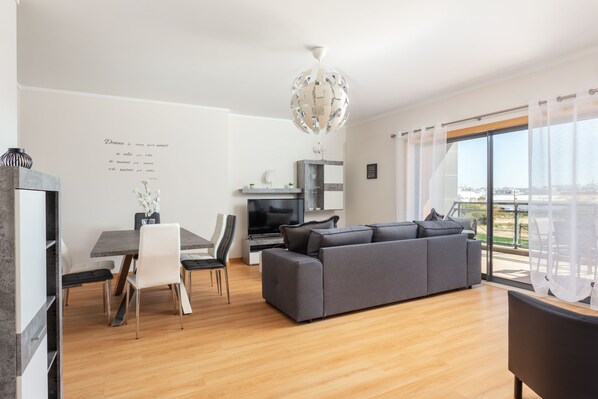 A modern living room with a grey sofa, dining table, and chairs. There is a TV, a decorative light fixture, and a view of the outdoors through large windows. - Luxury 2-Bedroom Apartment with Rooftop Pool, Village Marina, Olhão (Olhão)