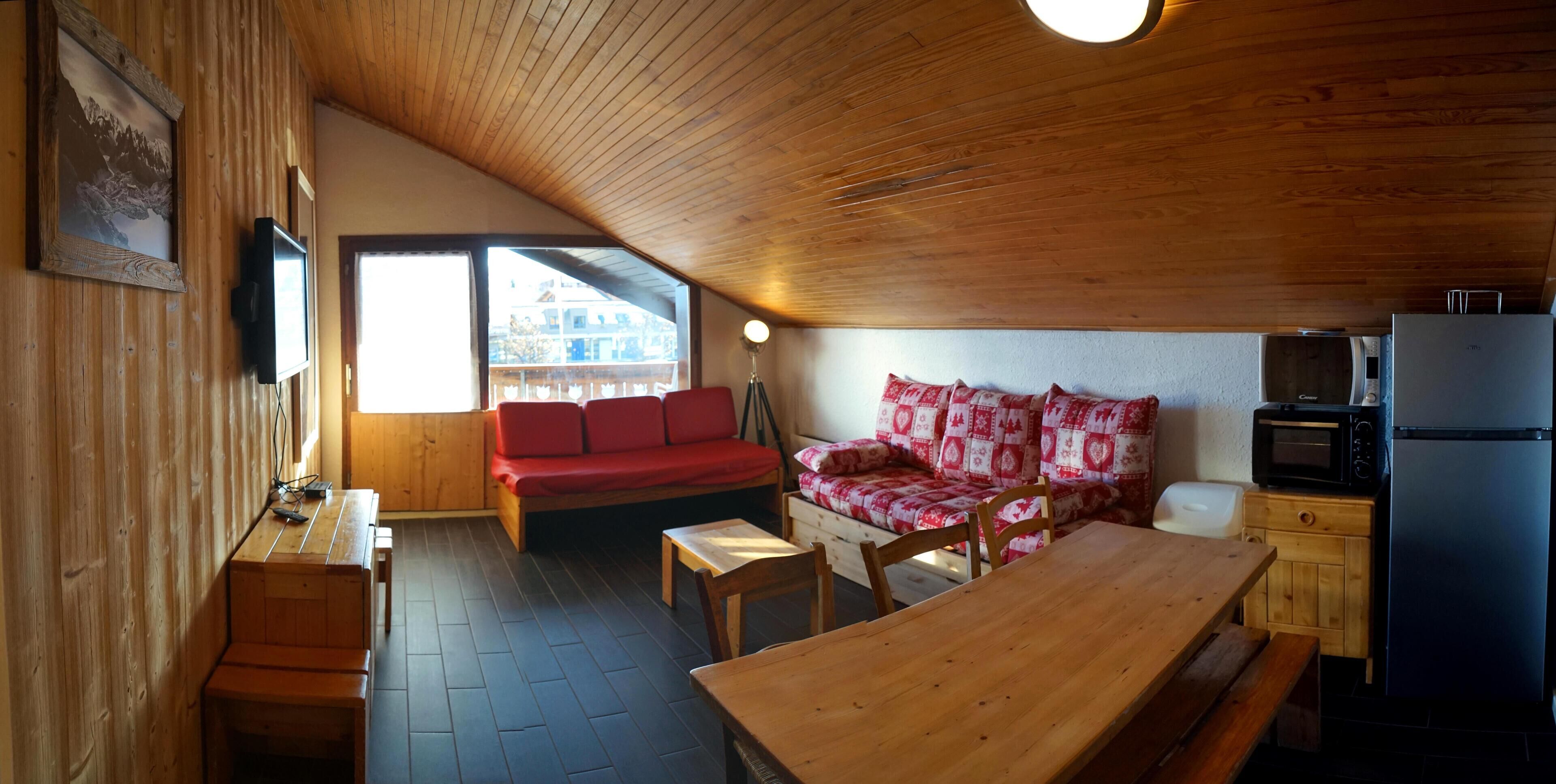A cozy cabin interior with a wooden ceiling, a red sofa, a wooden table, and a kitchen area with a refrigerator and microwave.