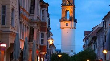 Una calle empedrada con edificios históricos, una torre del reloj y zonas de estar al aire libre.