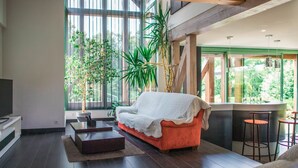 Un salon moderne avec un canapé rouge, une table basse et un téléviseur posé sur un meuble blanc. De grandes fenêtres offrent une vue sur la verdure environnante.