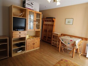 Um espaço de estar compacto com um móvel de entretenimento em madeira, uma área de refeições com mesa e cadeiras e uma pequena cozinha compacta.