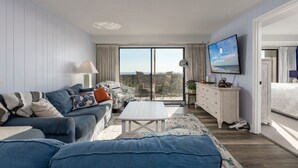 A modern living room with a sofa set, a coffee table, a TV mounted on the wall, and a view of the outdoors through large windows. - Direct oceanfront in convenient mid town location (Ocean City)