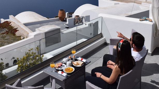 A terrace with a table set for two, overlooking a scenic view of the sea and a cliff. - Vitsentzos Suites (Santorini)