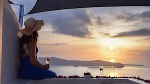A woman in a blue dress sitting by a pool at sunset with a glass of wine, a rose, and a hat on the table. - Vitsentzos Suites (Santorini)