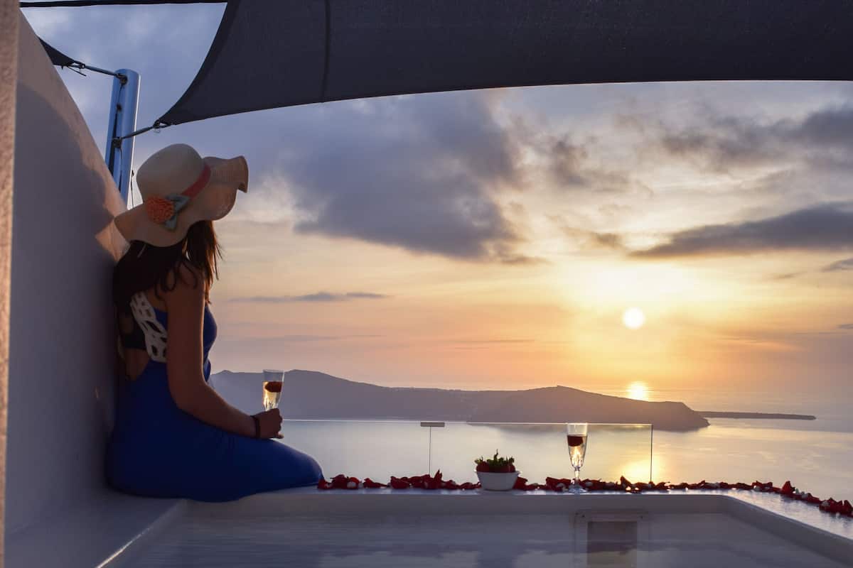 A woman in a blue dress sitting by a pool at sunset with a glass of wine, a rose, and a hat on the table.