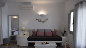 A modern living room with a sofa, a coffee table with decorative items, a wall-mounted light fixture, and a small plant. - Vitsentzos Suites (Santorini)