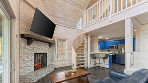 A spacious living area with a fireplace, a wooden staircase, a kitchen with blue cabinets, and a flat-screen TV mounted on the wall. - Top of the Line Treehouse Cabin (Cleveland)