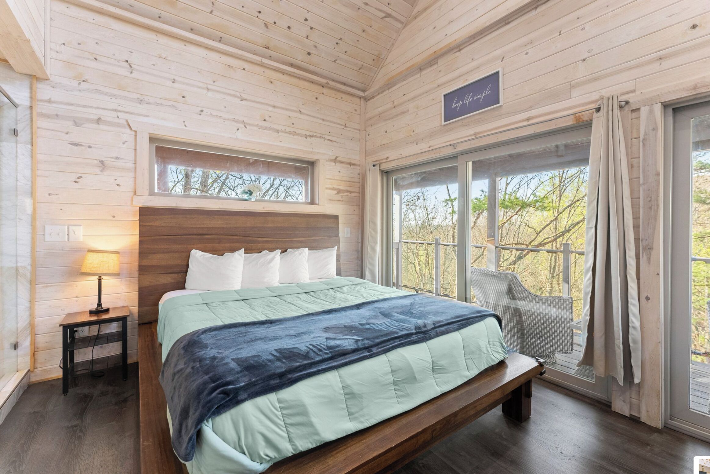 A wooden cabin bedroom with a bed, a nightstand, a window with curtains, and a view of trees.