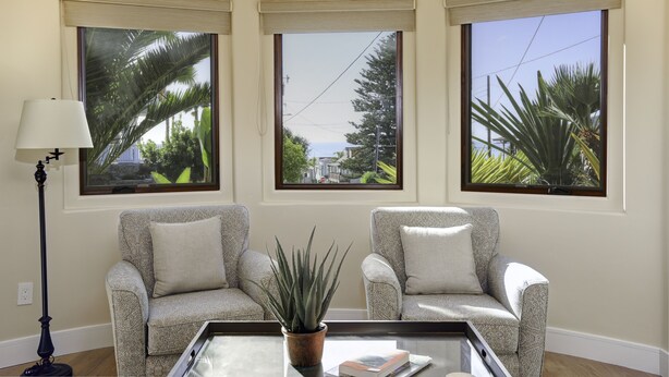 Un salón con tres ventanas amplias, dos sillones, una lámpara de pie y una mesa de centro con una planta y libros.