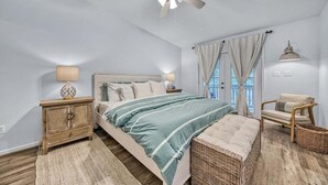 Un dormitorio con una cama, mesitas de noche, una silla, ventilador de techo y una ventana con cortinas.