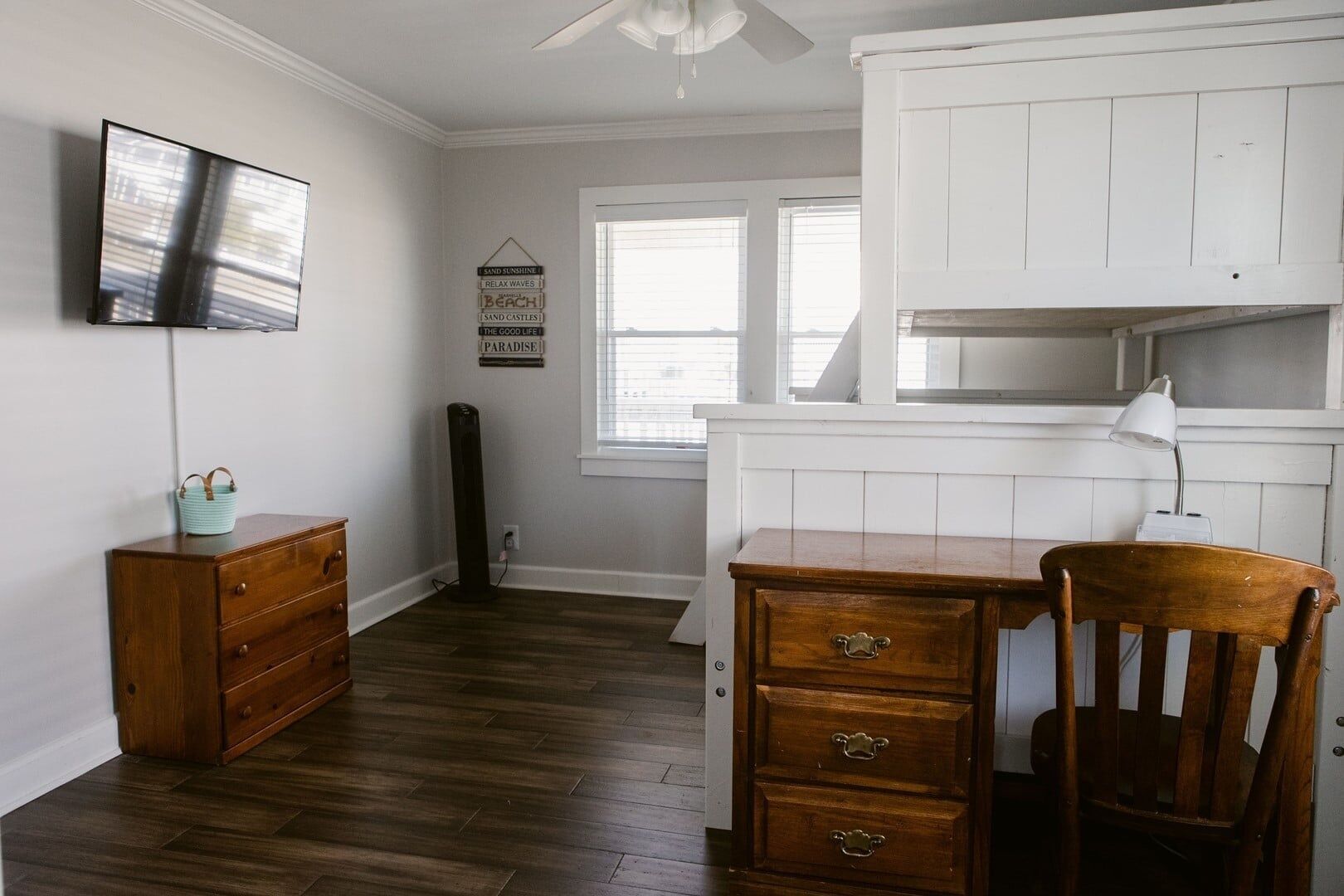Une pièce comprenant une commode en bois, une chaise, un bureau, un téléviseur et une fenêtre avec des stores.