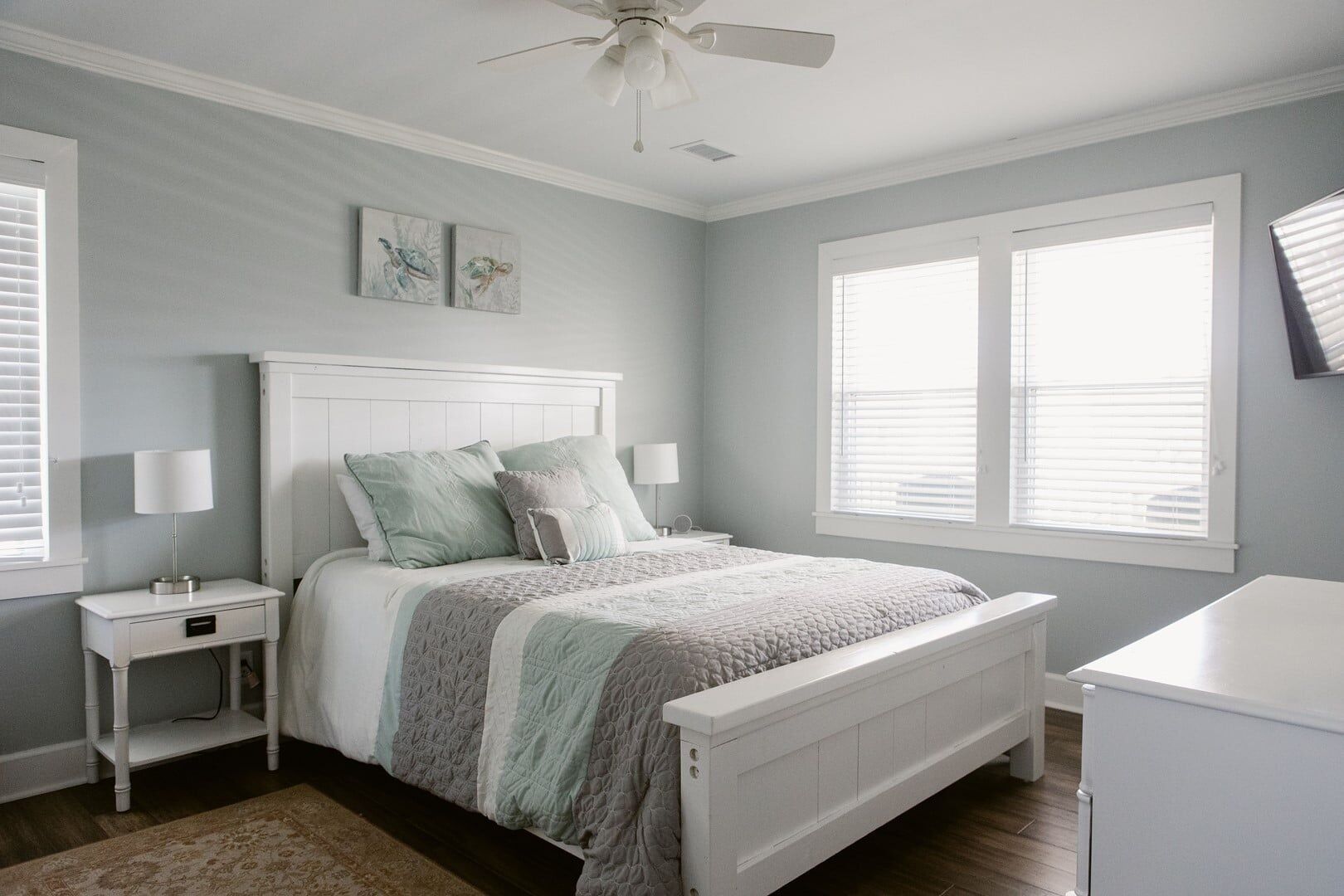A bedroom with a white headboard, a bed with a grey and white quilt, two bedside tables with lamps, and a window with blinds.