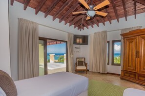 A bedroom with a bed, a wooden wardrobe, a chair, a television, and a ceiling fan. - Pristine Bay 1108 4 Bedroom Home by RedAwning (Roatan)