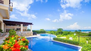 A swimming pool with a view of the ocean, surrounded by greenery and a building with a covered patio.