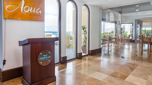 A hotel lobby with a wooden reception desk, a sign reading "Aqua by Pastina Bay," and a view of the outdoors through large glass doors.