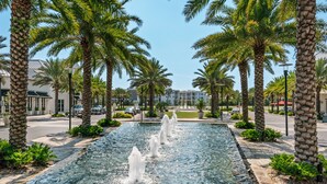 Une fontaine avec des jets d’eau au centre, entourée de palmiers et bordée d’une allée pavée.
