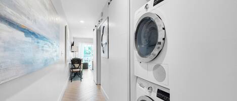 A laundry room with a washing machine and dryer, a painting on the wall, and a dining area in the background.