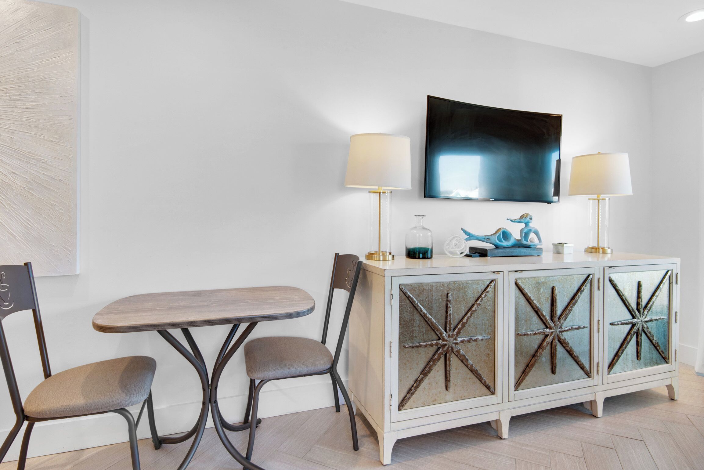 Un salon moderne avec une table en bois et deux chaises, un meuble blanc aux portes en verre décoratives et un téléviseur à écran plat fixé au mur.