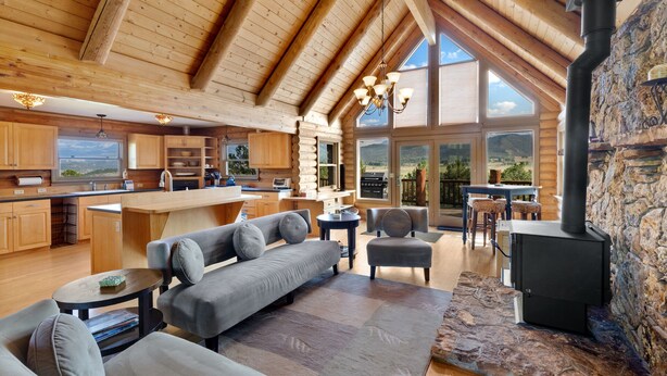 Eine geräumige Blockhütte mit hohen Decken, einem Steinkamin, einer Küche mit großen Fenstern und einem gemütlichen Wohnbereich mit Sofa und Sesseln.