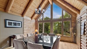 Ein Holzblockhaus-Interieur mit einem Esstisch, Stühlen, einer Chandelieber und großen Fenstern mit Blick auf Bäume.