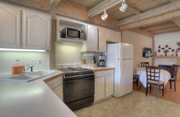 Una cocina con armarios blancos, estufa, microondas y refrigerador. Hay una mesa de comedor con sillas y una cafetera.