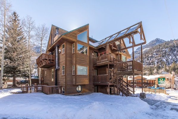 A multi-story wooden building with balconies and large windows, surrounded by snow and trees.