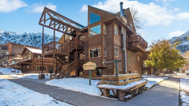 Un edificio de estilo cabaña de madera con una escalera que lleva a un balcón, un letrero que dice "Alpenglow Condominiums" y un banco cubierto de nieve.