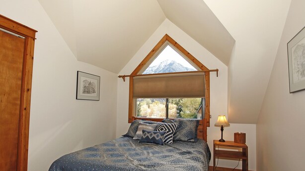 A bedroom with a sloped ceiling, a bed with patterned bedding, a bedside table with a lamp, and a window with a view of trees and mountains. - Next to Ouray Hot Springs | Walk to Main Street | Deck (Ouray)