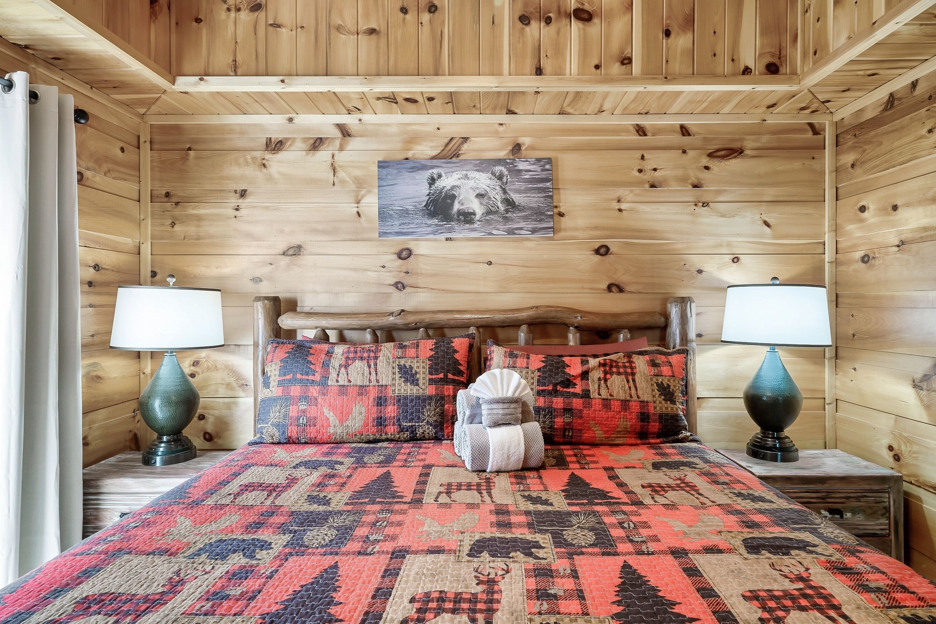 A wooden cabin bedroom with a bed, two nightstands, a lamp, and a bear-themed wall hanging.