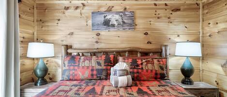 A wooden cabin bedroom with a bed, two nightstands, a lamp, and a bear-themed wall hanging.