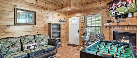 A cozy room with a wooden ceiling, a leather sofa with a bear-themed cushion, a foosball table, and a television displaying a football game.