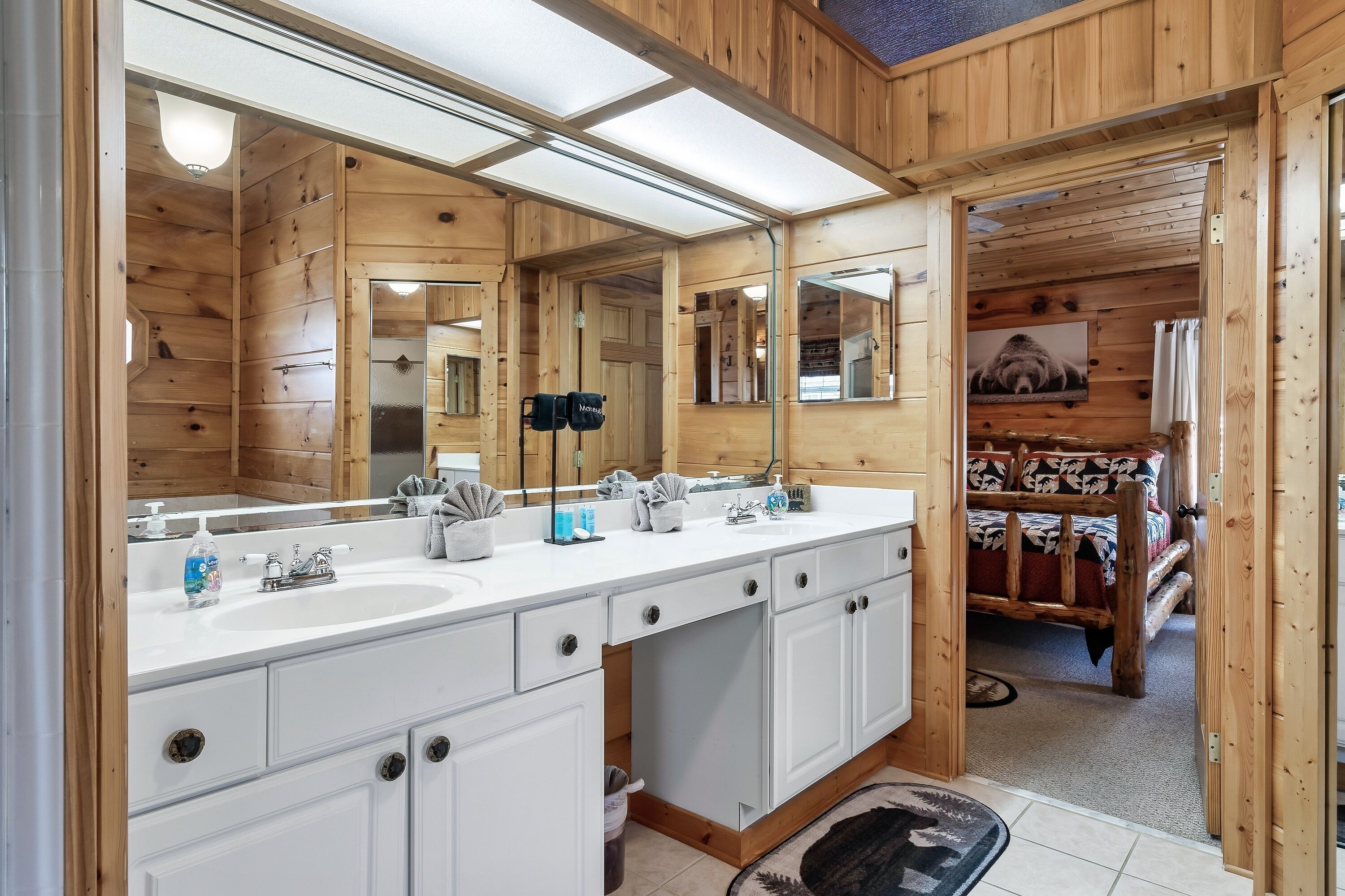 A wooden cabin-style bathroom with two sinks, a large mirror, and a bed visible through an open door.