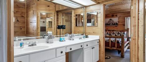 A wooden cabin-style bathroom with two sinks, a large mirror, and a bed visible through an open door.