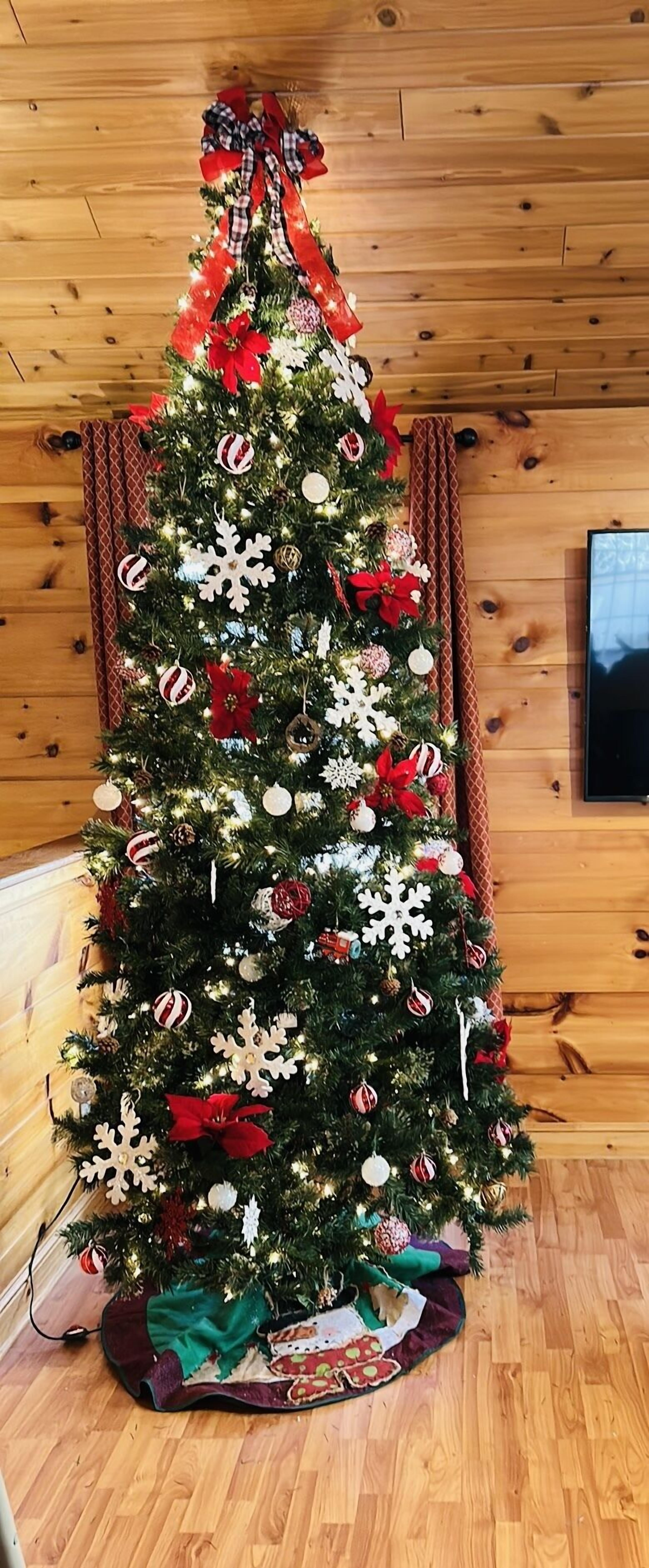 Een kerstboom versierd met decoraties en lichtjes, geplaatst in een houten kamer met een televisie aan de muur.