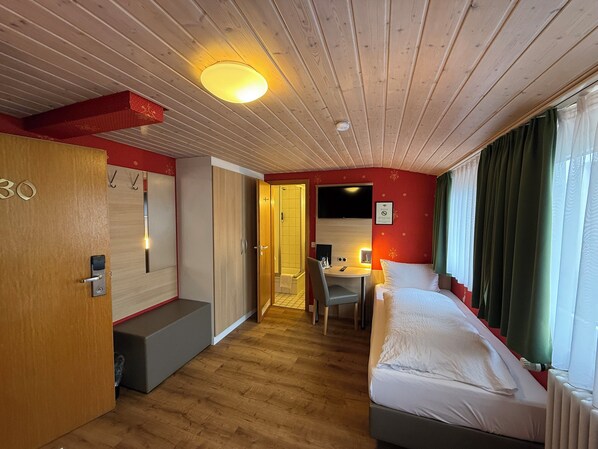 Habitació d'hotel amb sostre de fusta, un llit, escriptori amb cadira, banyet petit i finestra amb cortines.