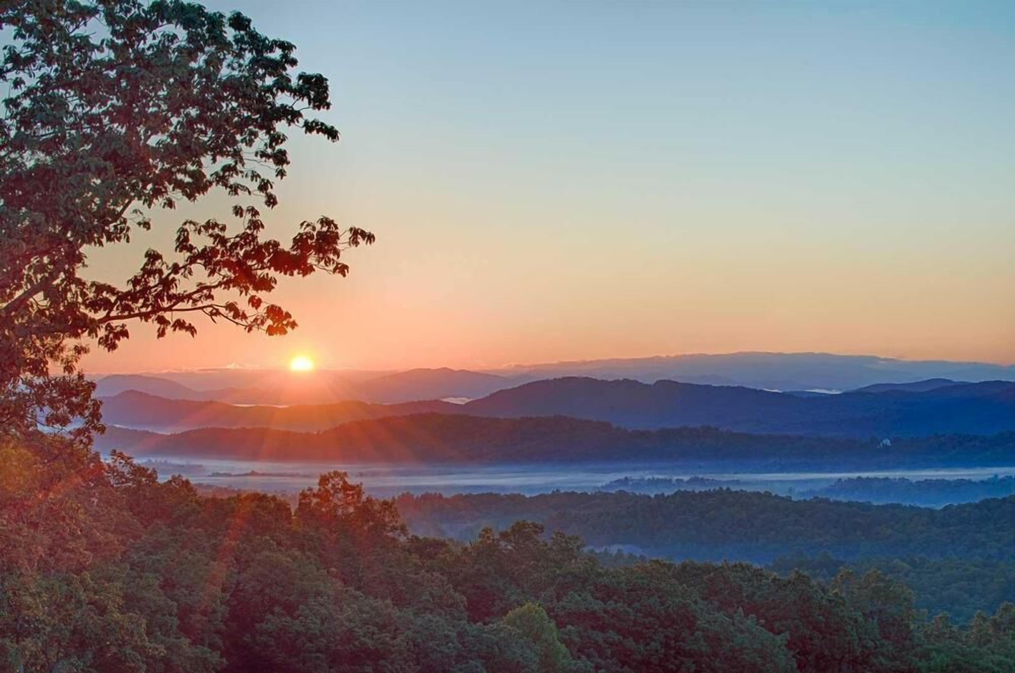 Sonnenaufgang über einer gebirgigen Landschaft mit dichtem Wald im Vordergrund.