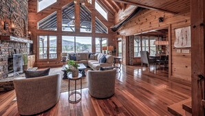 A spacious living room with a stone fireplace, large windows, and wooden beams.