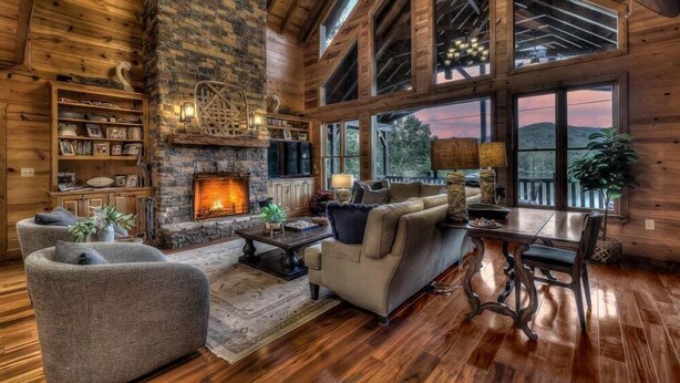 A cozy living room with a stone fireplace, wooden walls, and large windows offering a view of the outdoors.