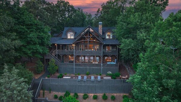 A large, multi-story log cabin with a balcony on each floor, surrounded by dense forest.