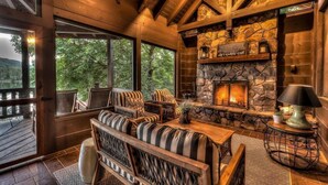 A cozy cabin interior with a stone fireplace, wooden furniture, and large windows overlooking a forested area.
