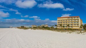 A beachfront hotel with multiple floors, a sandy beach in the foreground, and a clear blue sky with clouds. - Beachfront Condo Across from John's Pass -Madeira Norte- Gorgeous sunset views! (Madeira Beach)