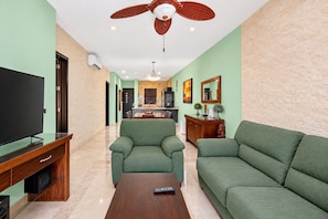 A living room with a flat-screen TV, green sofas, a coffee table, and a ceiling fan.