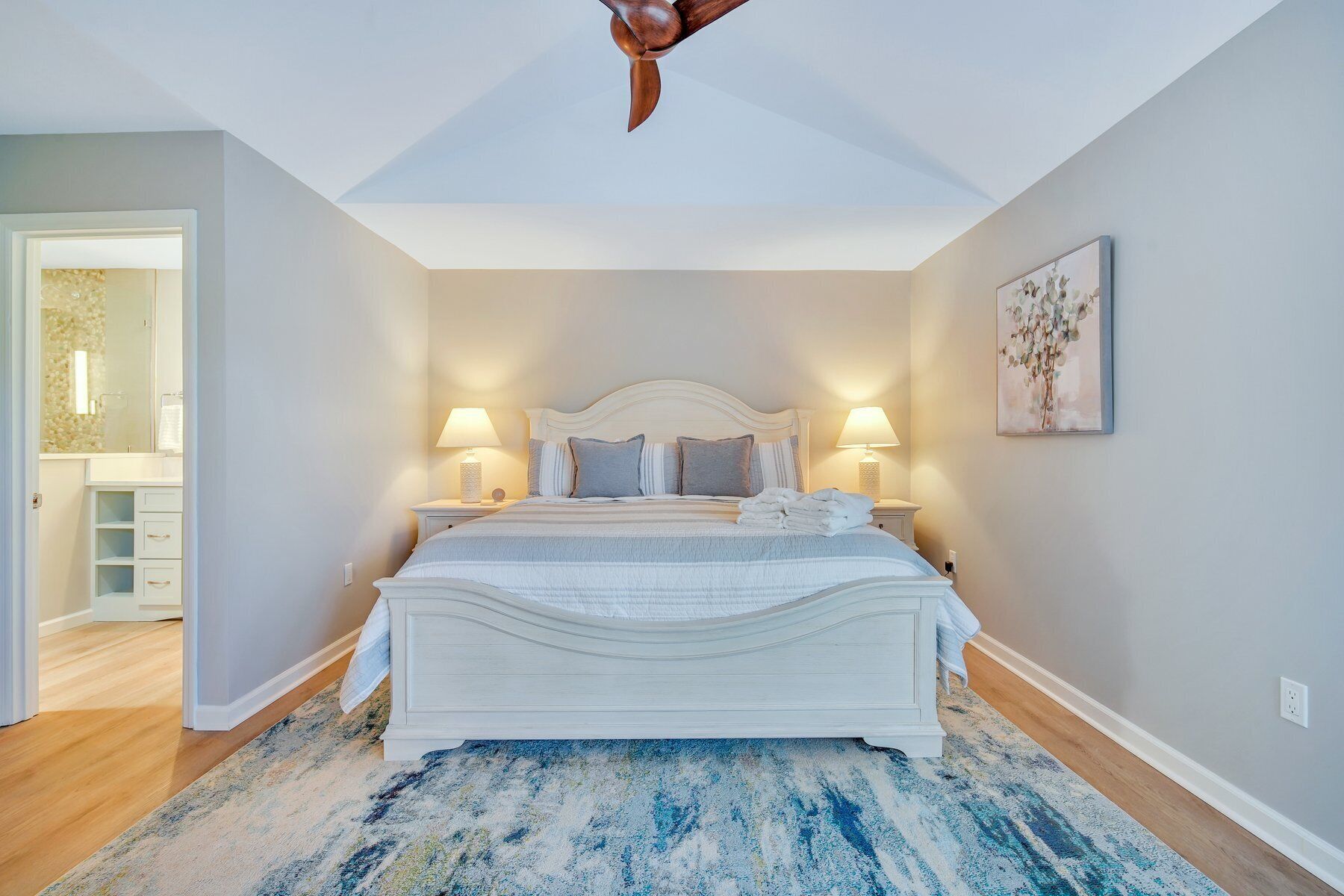 A bedroom with a large bed, two bedside tables with lamps, a ceiling fan, a painting on the wall, and a view into the bathroom.