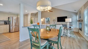 A modern kitchen with a stainless steel refrigerator, a dining area with a wooden table and green chairs, and a large ceiling fan.