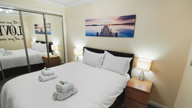 Une chambre d’hôtel avec un grand lit, deux tables de chevet avec des lampes, un miroir et un tableau représentant un coucher de soleil sur l’eau.