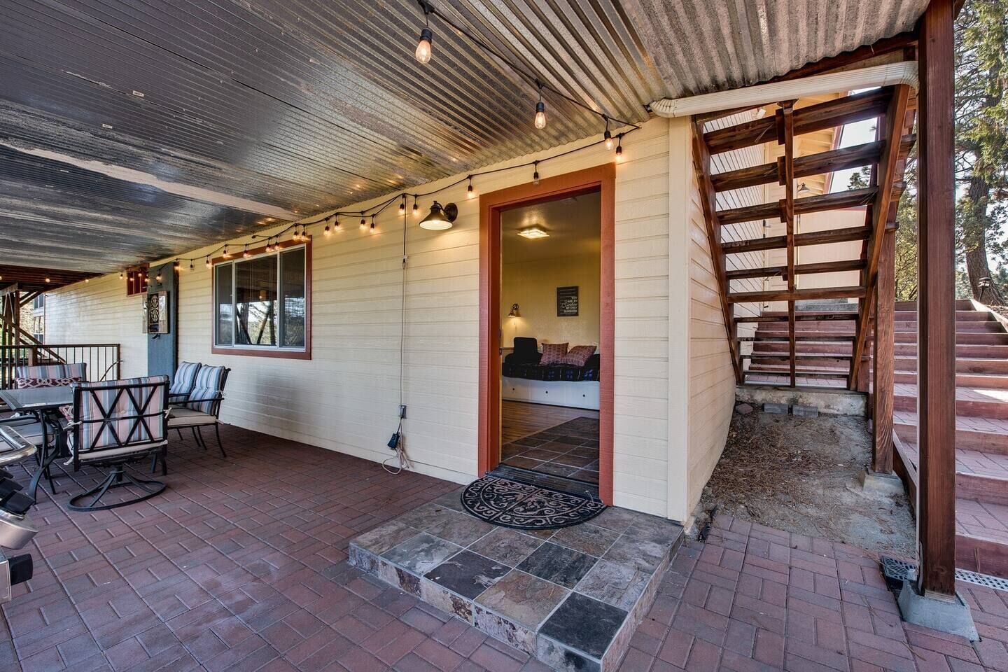 A covered patio area with outdoor seating, a wooden staircase, and a view of the interior.