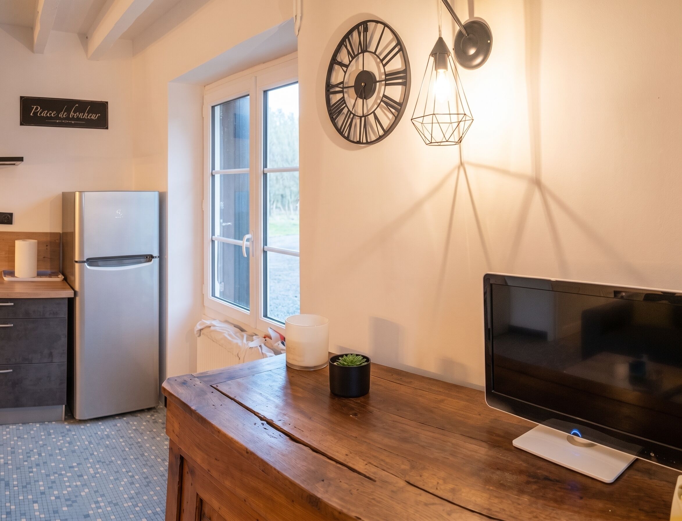 Une cuisine équipée d’un réfrigérateur, d’une fenêtre, d’une horloge murale, d’un luminaire suspendu et d’un téléviseur à écran plat.