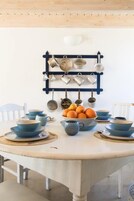 Un comedor con una mesa redonda dispuesta con vajilla azul y un frutero. Un perchero de pared exhibe diversos utensilios de cocina.
